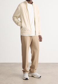 Man som bär en krämfärgad zip-up jacka, vit T-shirt, beige byxor och vita sneakers, står med en hand i fickan.