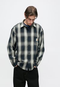 Mėlynos ir kreminės spalvų languota flanelinė marškinė, su mygtukais priekyje, ilgomis rankovėmis ir priekinėmis kišenėmis su logotipo pleistru.