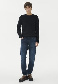 Jaunas vyras stovi su rankomis kišenėse, apsirengęs juodu megztiniu, mėlynais džinsais ir rudiem žygio batais ant paprasto balto fono.