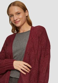 Burgunderfarbener Strickcardigan mit Zopfmuster, offener Vorderseite und gerippten Bündchen, getragen über einem grauen Oberteil. Weiche Textur, lässiges Design.
