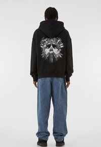 Sort hættetrøje med grafisk design på ryggen, der viser en central figur og blomsterakcenter. Bæres sammen med løse blå denimjeans.