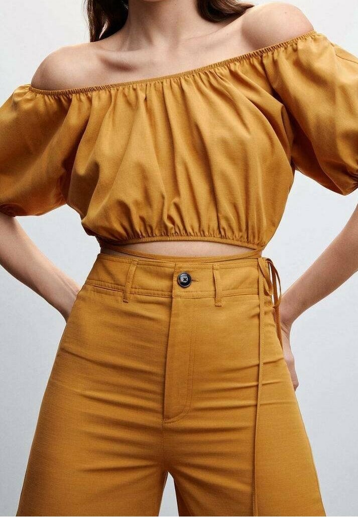 Femme portant un top court jaune moutarde à épaules dénudées avec manches bouffantes et un pantalon taille haute avec un bouton et un détail de lien sur le côté.