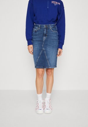 Jeans-Bleistiftrock in mittlerem Blau mit einer geschwungenen vorderen Naht. Kombiniert mit einem blauen Sweatshirt mit Reißverschluss und weißen Sneakers.