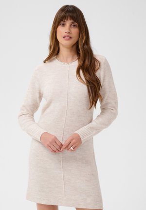 Donna con capelli lunghi castani che indossa un vestito a maglia beige chiaro a maniche lunghe, in piedi con le mani giunte davanti, sfondo neutro.