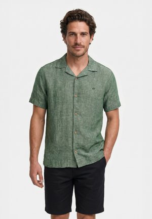 Homme portant une chemise verte à manches courtes boutonnée et un short noir, debout avec une main dans la poche sur un fond blanc uni.