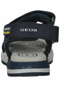 Sandalias azules y negras con parte superior texturizada, correa ajustable, suela de goma y entresuela blanca. Presenta el logotipo "GEOX" destacado.