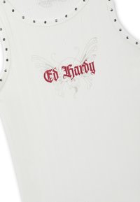 White ribbed tank top with metal eyelets on shoulder edges and red "Ed Hardy" text over faded butterfly graphic at center chest.