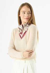 Foulard en soie à base crème avec des motifs abstraits rouges et noirs, soigneusement plié en forme de triangle, porté sur un pull léger.