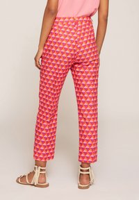 Kvinna som bär rosa geometriskt mönstrade cropped byxor och beige sandaler med fransar, stående framför en enkel bakgrund.