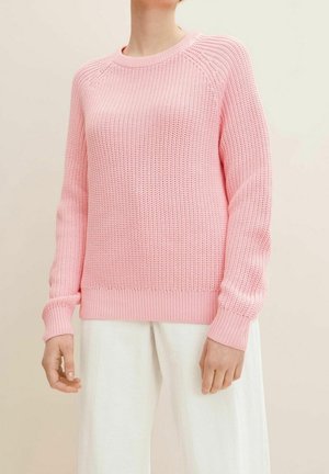 Personne portant un pull rose texturé à manches longues associé à un pantalon blanc ample, sur un fond clair uni.