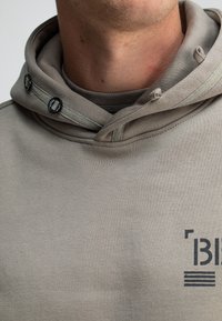 Beige hoodie van een gestructureerde stof, met een trekkoordkap, twee knopen en een bedrukt marineblauw logo op de voorkant.