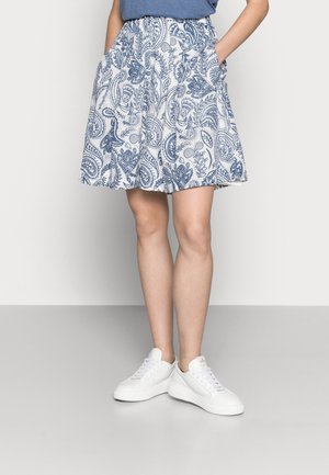 Jupe mi-longue aux genoux à motif cachemire bleu et blanc, portée avec des baskets blanches et un haut bleu rentré, mains dans les poches.