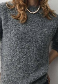 Pull gris en maille texturée avec un col rond et des manches trois-quarts, accompagné d'un collier de perles. Matière douce et confortable.