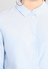 Kvinna klädd i en ljusblå knappad skjorta med krage och en stor guldfärgad hoopsörhänge synlig på vänster sida.