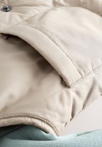 Tissu de veste matelassée beige avec coutures visibles, fermeture à boutons et col plié, superposé sur un vêtement bleu clair texturé.