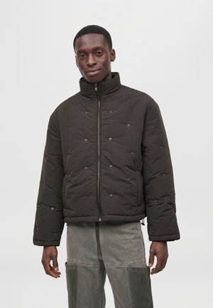 Un homme porte une veste matelassée foncée à col montant et à fermeture éclair sur le devant, avec un pantalon gris, sur un fond uni.