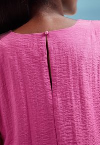 Pink, textured garment with a rear keyhole opening secured by a small button. Soft fabric and relaxed silhouette evident.