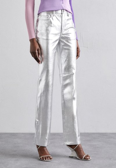 Pantalons métalliques argentés à jambes droites avec une surface brillante, poches avant et fermeture à bouton. Associés à des sandales blanches à lanières.