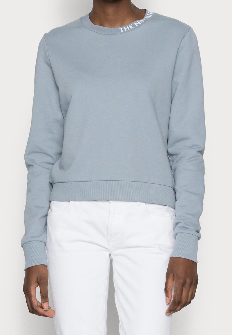 Personne portant un sweat-shirt bleu clair à col rond avec du texte blanc sur le col et un pantalon blanc, debout devant un fond uni.