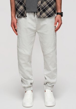 Man draagt lichtgrijze joggerbroek, witte sneakers, een grijs shirt en een open geruit overhemd met een horloge aan de linkerpols.
