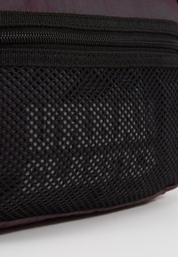 Gros plan sur une poche en mesh noir et une fermeture éclair sur un tissu violet foncé, probablement une partie d'un sac ou d'un sac à dos.