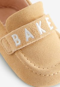 Kerge suède mokassini stiilis kingad, millel on pehme tekstuur ja ees valge broderiga "BAKE" rihm. Nähtavad õmblused.