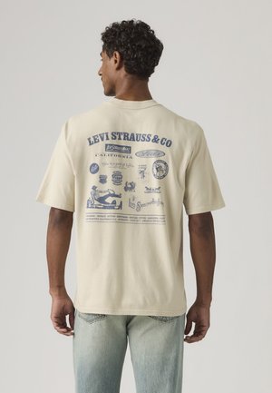 Hombre visto desde atrás con una camiseta beige de Levi's con logos y texto vintage en azul, combinado con jeans azul claro.