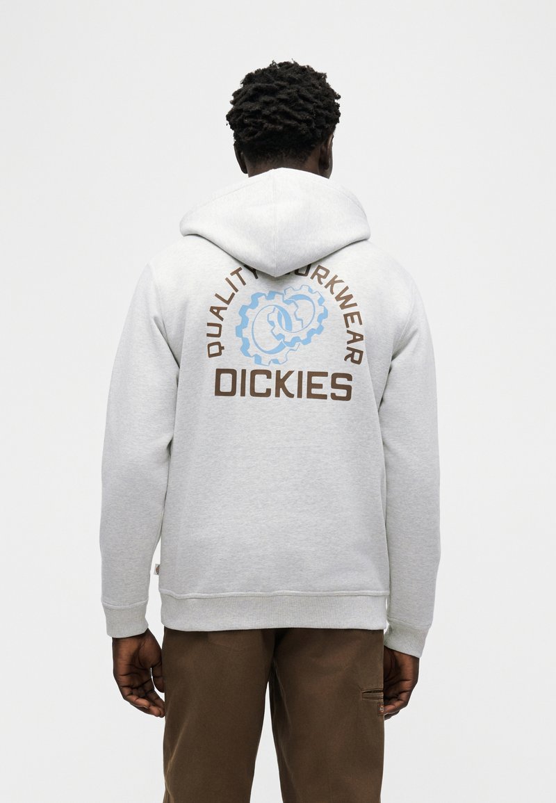 Uomo che indossa una felpa con cappuccio Dickies grigio chiaro con logo gear blu e marrone e la scritta "Quality Workwear" sul retro, abbinata a pantaloni marroni.