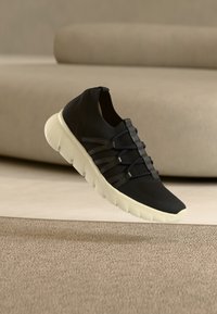 Μαύρο αθλητικό παπούτσι slip-on με λευκή χοντρή σόλα, ελαστικά κορδόνια και πλεκτό επάνω μέρος, αιωρούμενο πάνω από μπεζ χαλί με μπεζ καναπέ στο φόντο.