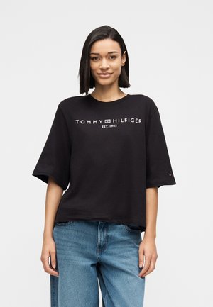 Frau mit glatten schwarzen Haaren, die ein schwarzes Tommy Hilfiger T-Shirt und blaue Jeans trägt, vor einem schlichten weißen Hintergrund stehend.