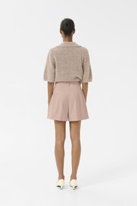 Beige gebreide trui met een kraag, gecombineerd met lichtroze geplooide shorts. Model draagt crèmekleurige puntige schoenen, haar in een knot gestyled.