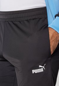 Pantaloni sportivi negri, cu o textură netedă, având un logo alb Puma pe partea stângă și un talie elastică.