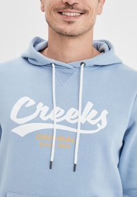 Felpa di un blu chiaro con cappuccio regolabile, caratterizzata dalla scritta bianca in grassetto "Creeks" e "ORIGINAL Since 1974" in arancione stampata sul davanti. Tessuto morbido.