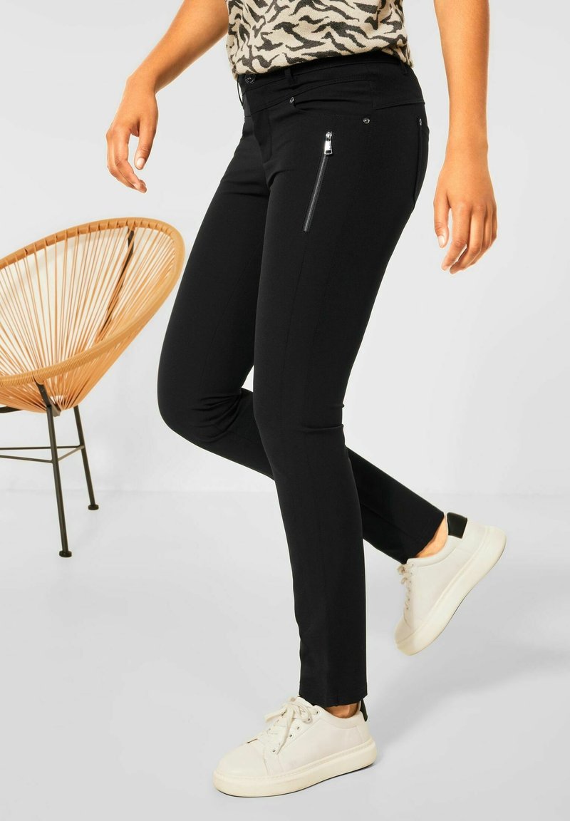 Black trousers featuring a slim fit, zippered side pocket, and a smooth texture. Paired with white sneakers and a patterned top.