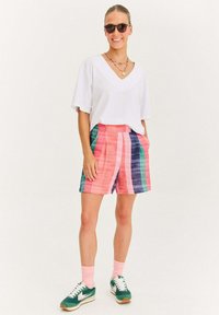 Žena v černých slunečních brýlích, bílé tričko s V výstřihem, barevné proužkované šortky, růžové ponožky a zelené tenisky, usmívající se s rukama v kapsách.