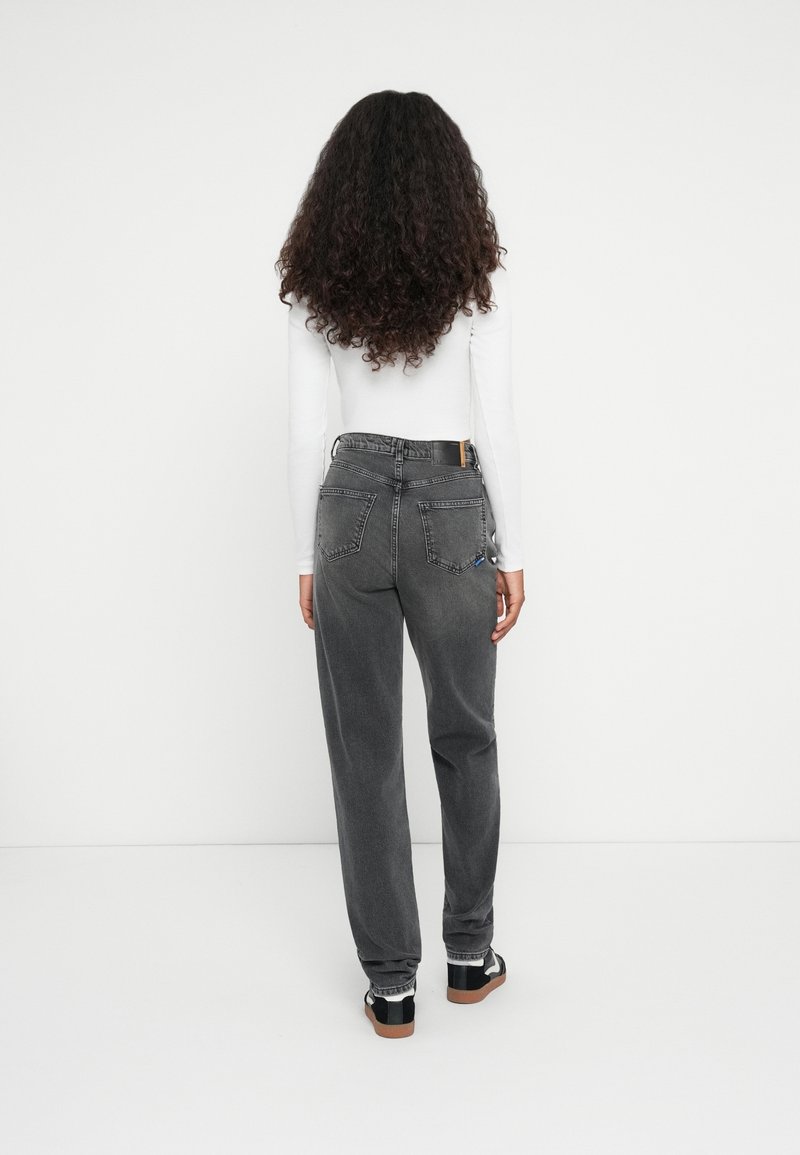 Femme aux longs cheveux bouclés portant un haut blanc à manches longues, un jean noir délavé taille haute et des baskets noires, debout de dos face à l'avant.