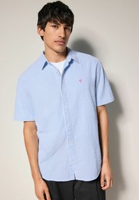 Camicia a maniche corte con bottoni di colore azzurro chiaro con righe bianche, dotata di un piccolo logo di una palma rosa e di un tessuto strutturato.