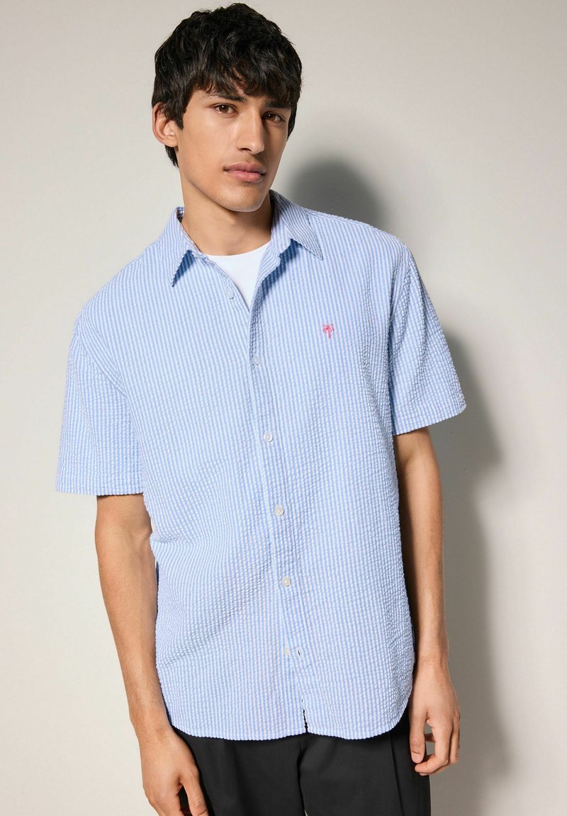 Camicia a maniche corte con bottoni di colore azzurro chiaro con righe bianche, dotata di un piccolo logo di una palma rosa e di un tessuto strutturato.