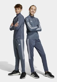 Graue Sportanzüge mit weißen Streifen, bestehend aus Reißverschlussjacken und schmal geschnittenen Hosen, kombiniert mit schwarzen Turnschuhen. Weiches, atmungsaktives Material.