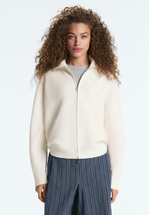 Frau mit lockigem Haar, die einen cremefarbenen, zum Reißverschluss geöffneten Pullover über einem grauen Shirt und blau gestreiften Hosen vor weißem Hintergrund trägt.