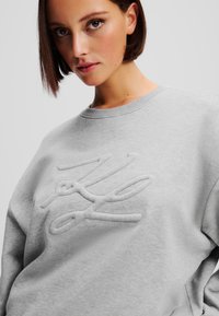 Hellgrauer Sweatshirt mit Rundhalsausschnitt, das in der Mitte ein strukturiertes, gesticktes Logo aufweist. Hergestellt aus weichem, bequemem Material.
