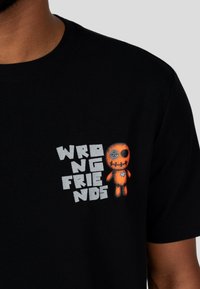 Černé tričko s potiskem, na kterém je text "WRONG FRIENDS" a oranžová panenka s vyšitými detaily a knoflíkovým okem.