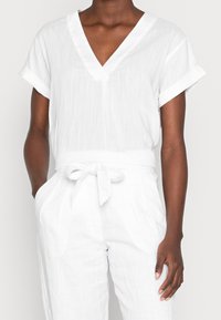 Top bianco a maniche corte e scollo a V con risvolti, realizzato in tessuto testurizzato; abbinato a pantaloni bianchi larghi con cintura annodata.