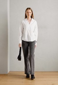 Hvit skjorte med krage, grå utsvingte bukser, svarte platåskjorter og en svart håndveske, satt mot en nøytral bakgrunn med tregulv.