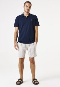 Donkerblauwe polo met korte mouwen, met een klein logo. Gecombineerd met lichtbeige shorts met verticale blauwe strepen, gedragen met zwarte sandalen.