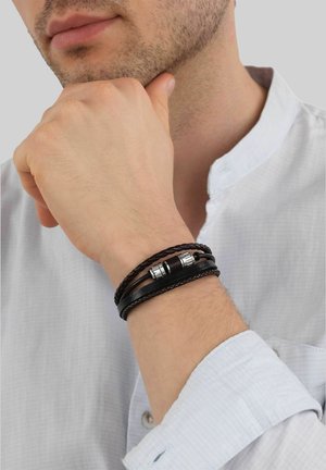 Hombre con camisa blanca y pulsera de cuero negra de varias capas en la muñeca, mano apoyada en la barbilla sobre fondo gris.