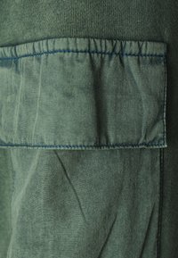 Tissu vert avec une surface texturée et une poche plaquée avec des coutures bleues. Le design comprend des sections lisses et froissées.