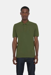 Polo verde realizzato in tessuto liscio, con un colletto, due bottoni, maniche corte e un design semplice, abbinato a jeans scuri.
