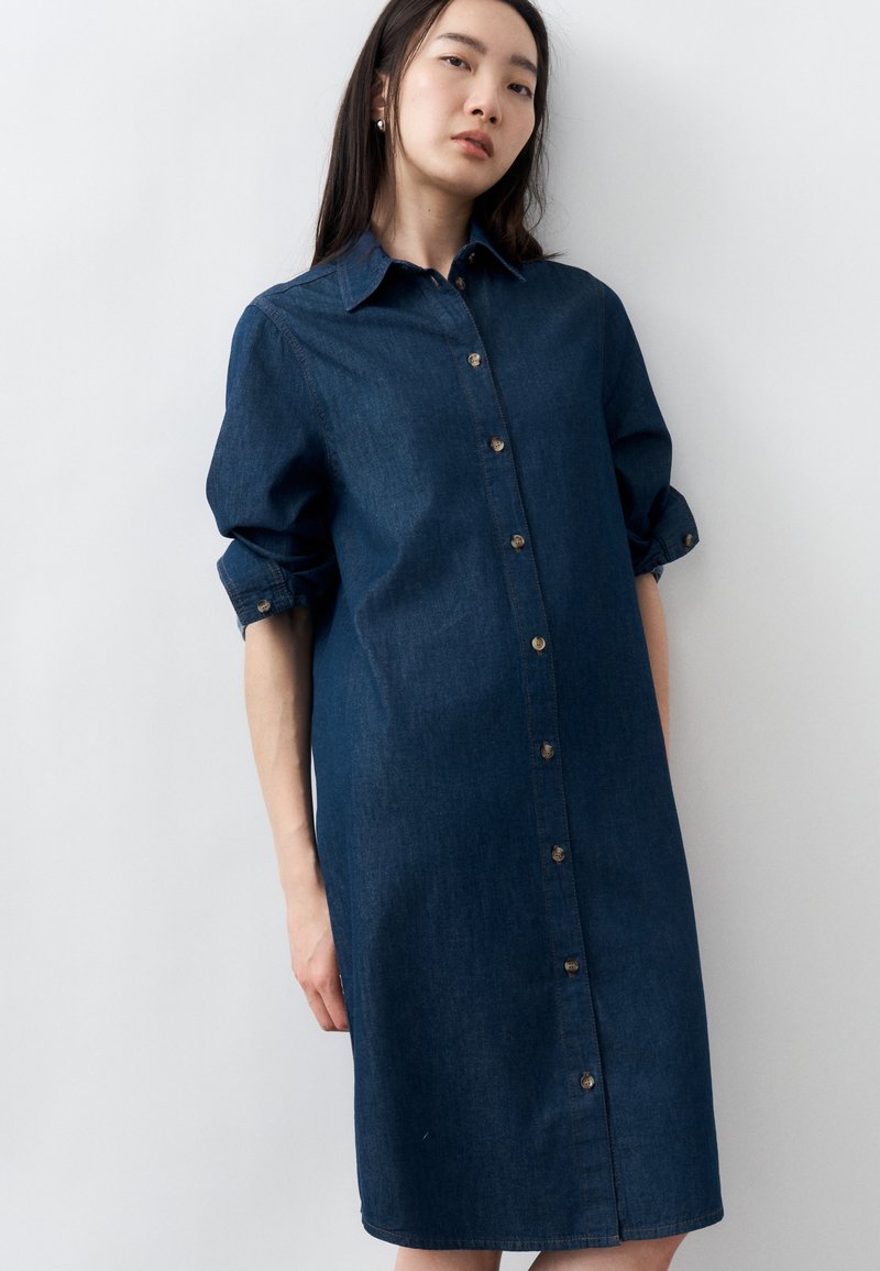 Naine, kes kannab pikkade varrukatega, nööpidega tumedast teksast kleiti, seisab valge tausta ees.