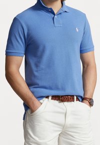 Polo shirt azzurro chiaro con finitura textured, colletto a due bottoni e piccolo emblema rosa. Indossato con pantaloni bianchi e una cintura intrecciata marrone.